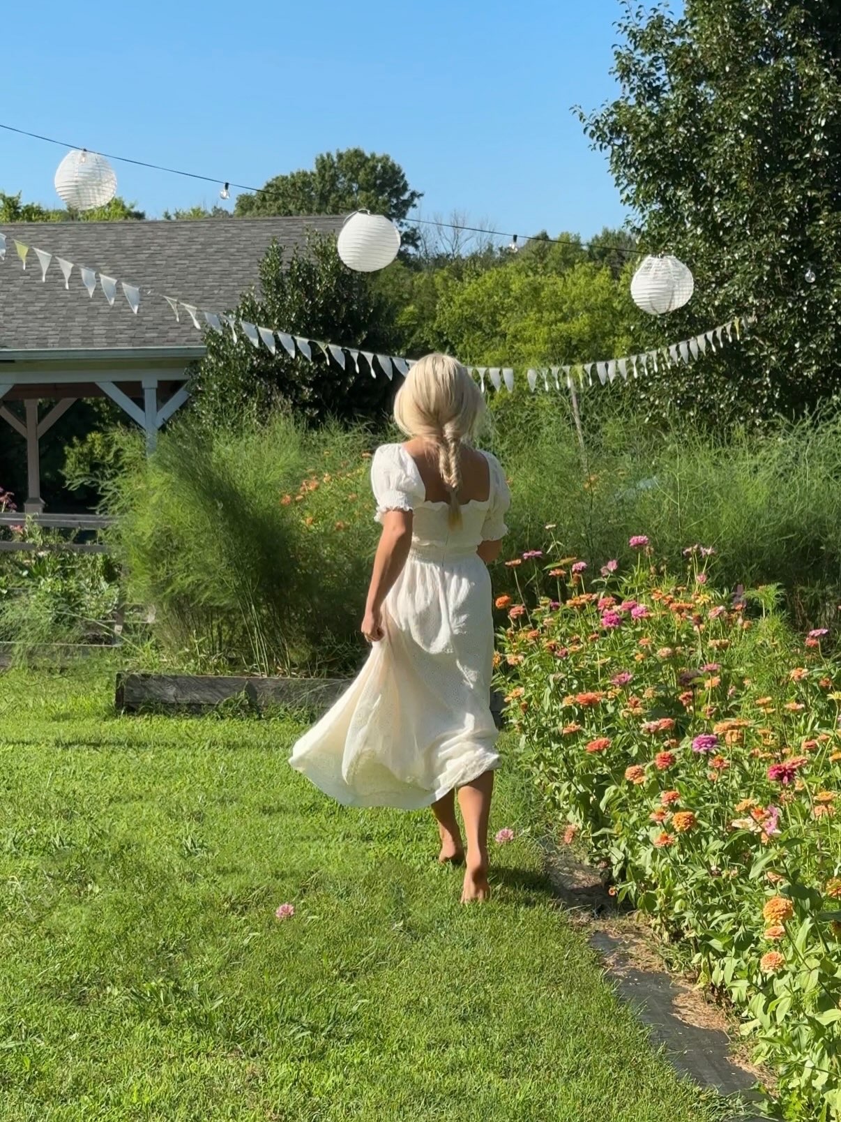 A woman in a white dress walks beside blooming zinnias in a sunny late-summer garden