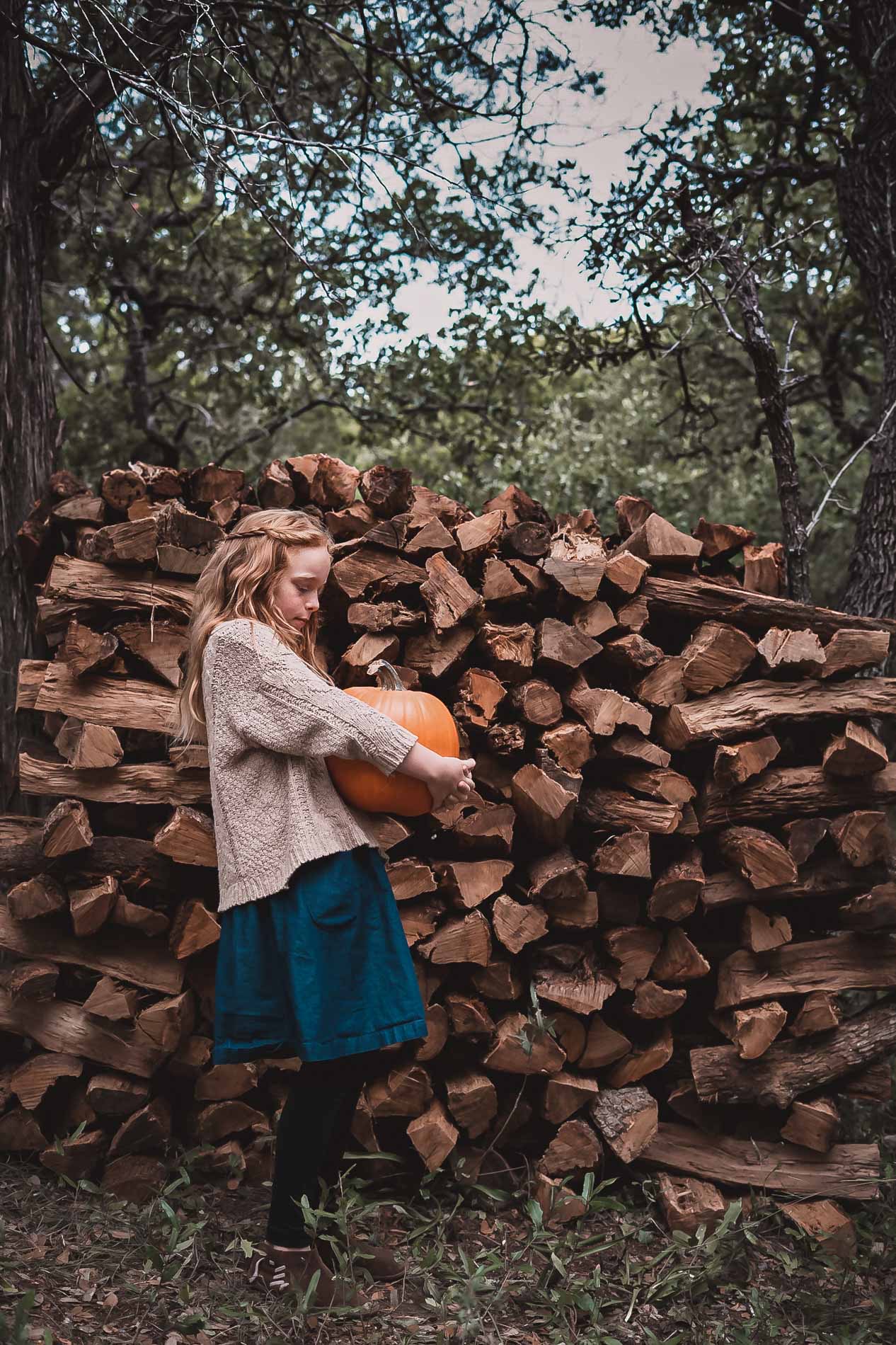 A child holding a pumpkin beside a stack of firewood at Frémont Farm